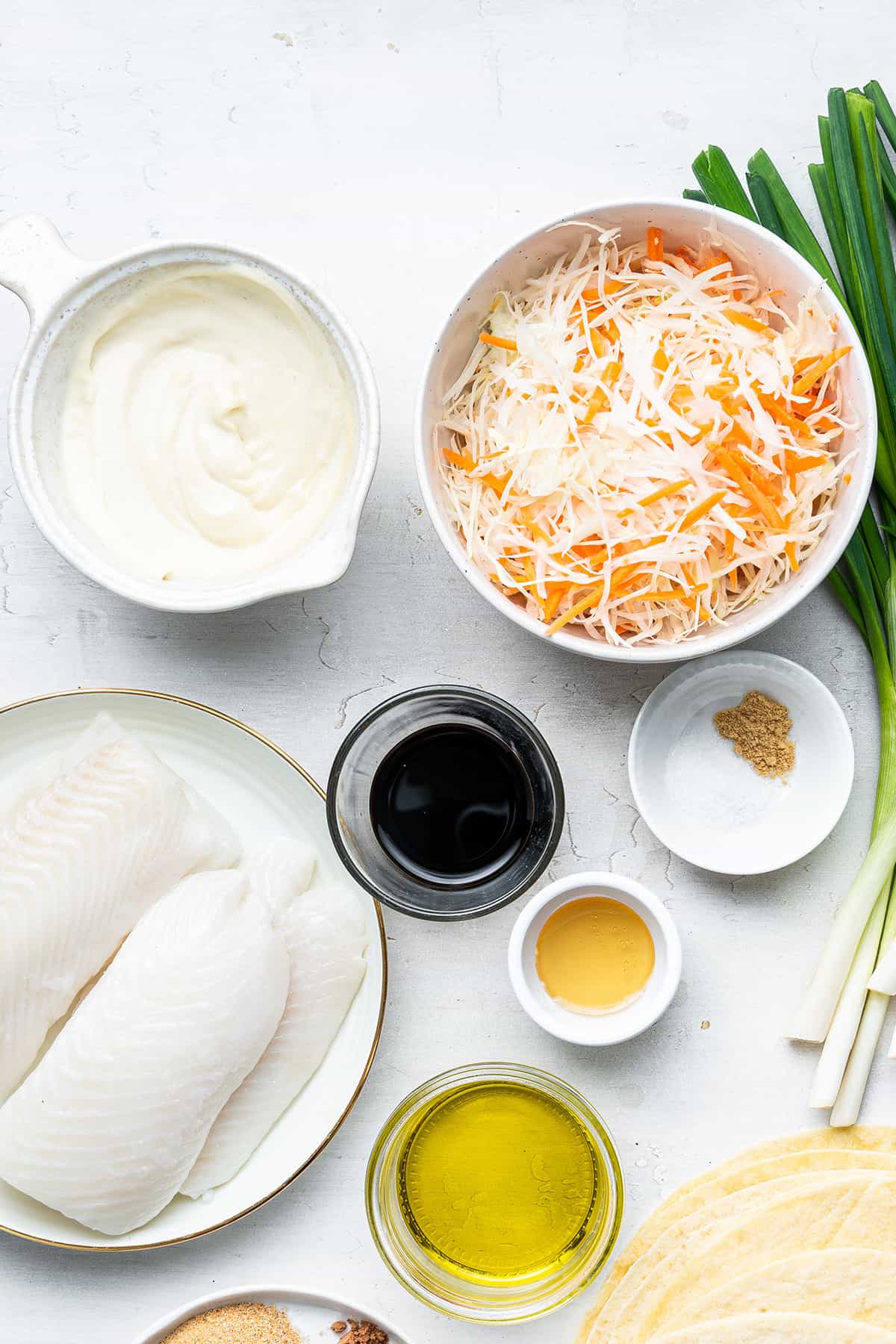 Overhead view of a bowl of carrots and cabbage, a bowl of mayonnaise, a bowl of soy sauce, a bowl of honey, a bowl of salt and ground ginger, a bowl of oil, a plate of raw fish, and some scallions