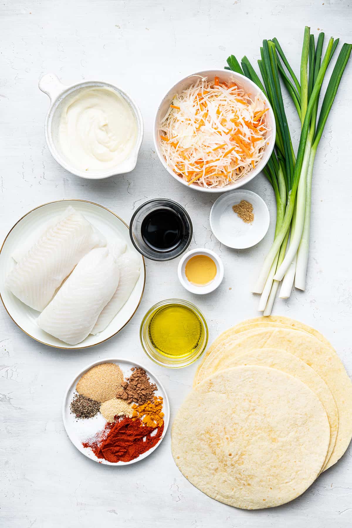 Overhead view of the ingredients for blackened fish tacos: a plate of mahi-mahi, a bowl of shredded carrots and cabbage, a bowl of dressing, a bowl of oil, a bowl of spices, some scallions, and a stack of tortillas
