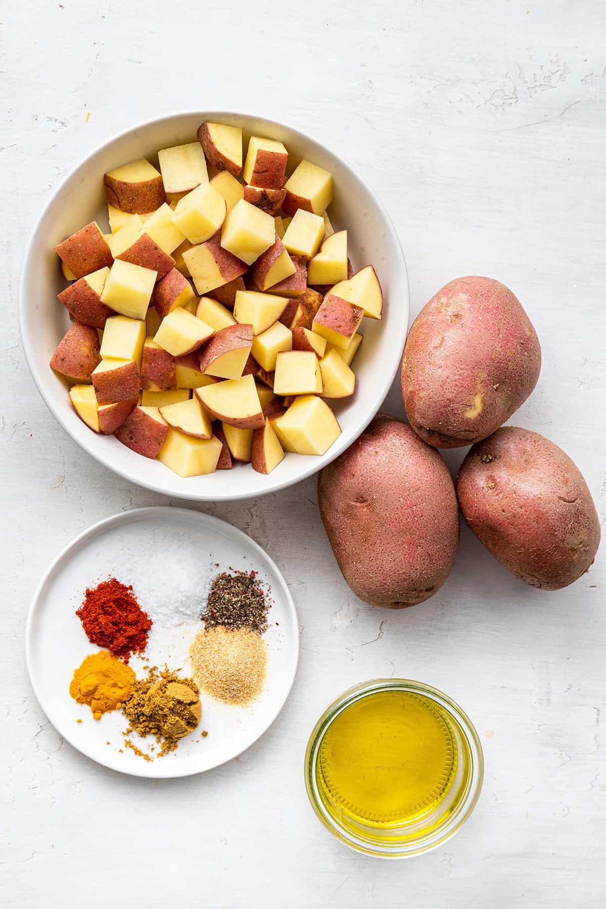 Overhead view of the ingredients needed for breakfast potatoes: three red potatoes, a bowl of chopped up potato cubes, a bowl of olive oil, and a plate with salt, pepper, garlic powder, smoked paprika, turmeric, and ras el hanout.