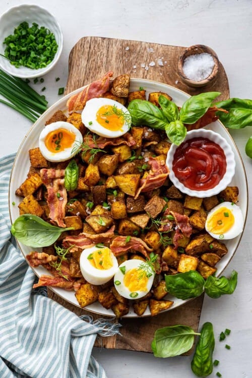 Overhead view of a plate of breakfast potatoes topped with bacon, basil, and soft boiled eggs, with a bowl of ketchup, a bowl of salt, and a bowl of chives