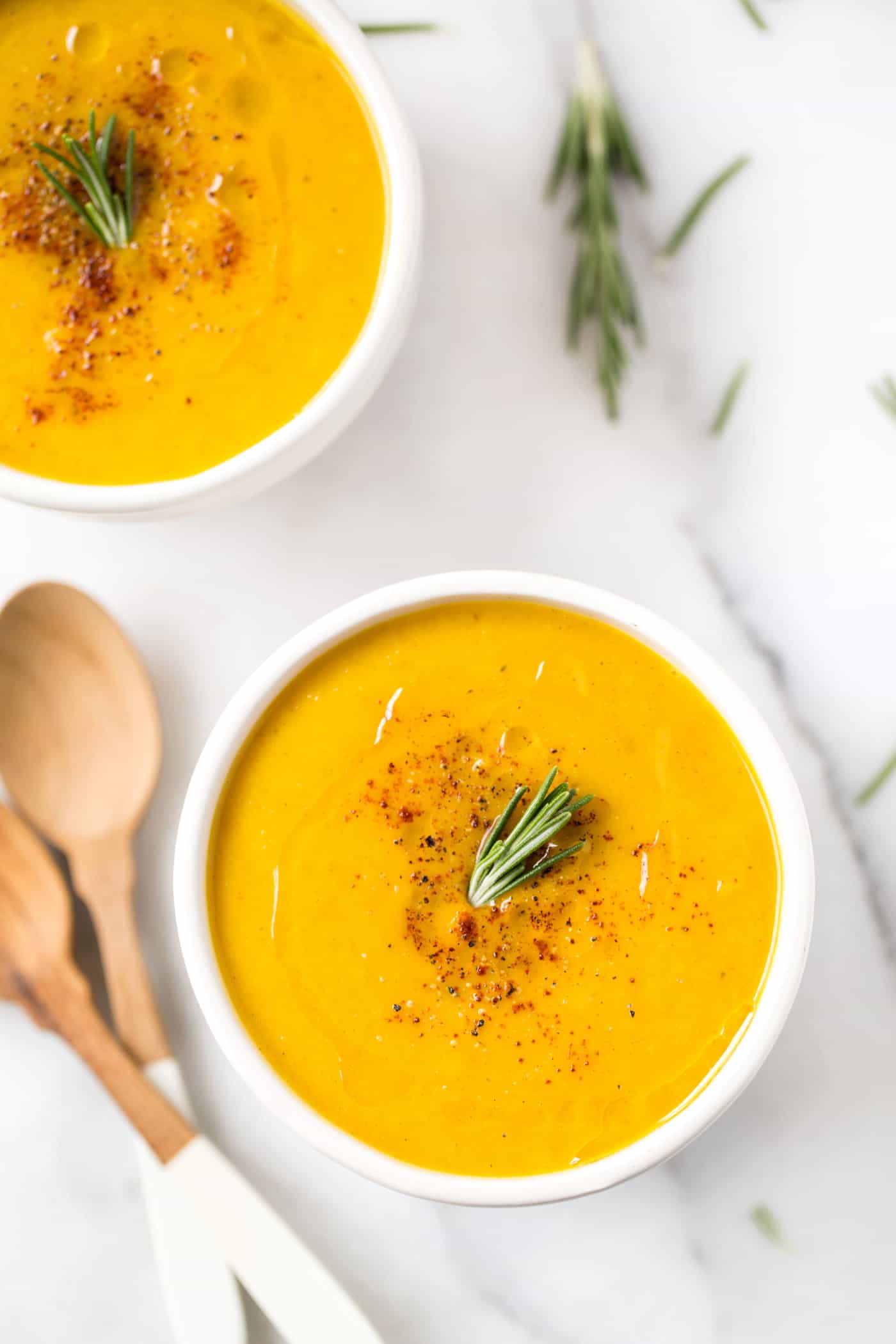 Two bowls of squash soup topped with olive oil, cracked black pepper, and fresh rosemary, with wooden spoons and rosemary in the background