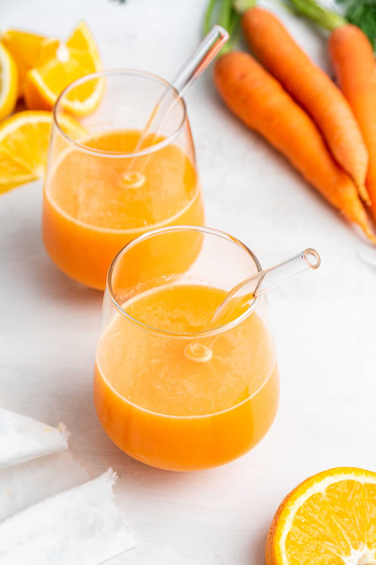 Two glasses of carrot juice with glass straws, surrounded by sliced oranges and carrots