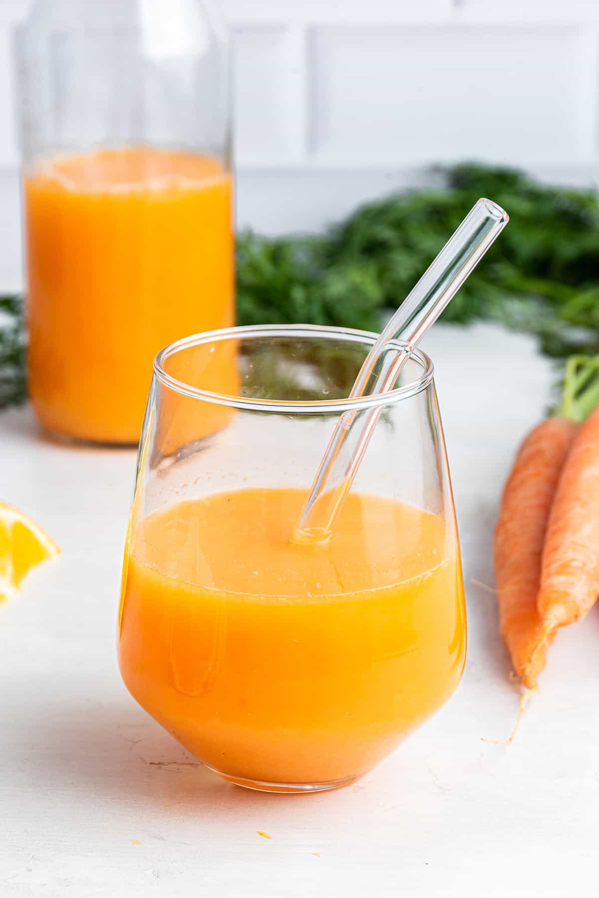 Glass of carrot juice with glass straw, with bottle of juice and bunch of carrots in background