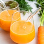 Two glasses of carrot juice with glass straws set next to carrot on counter