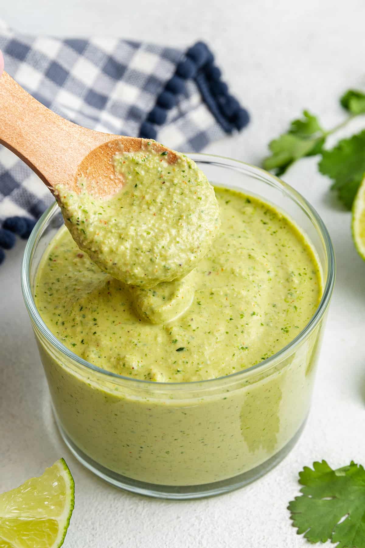 A jar of cilantro lime sauce with a spoon being removed from it, surrounded by lime slices, cilantro leaves, and a kitchen towel