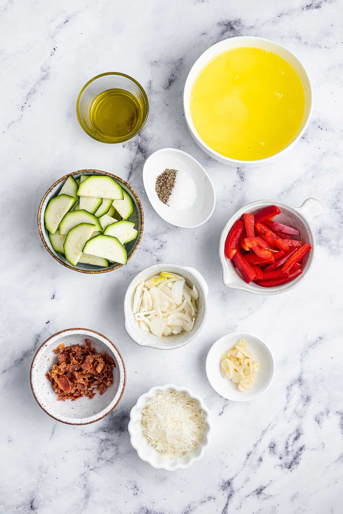 Overhead view of the ingredients needed to make egg white omelets: a bowl of egg whites, a bowl of oil, a bowl of salt and pepper, a bowl of onions, a bowl of garlic, a bowl of bell peppers, a bowl of zucchini, a bowl of bacon, and a bowl of parmesan cheese.