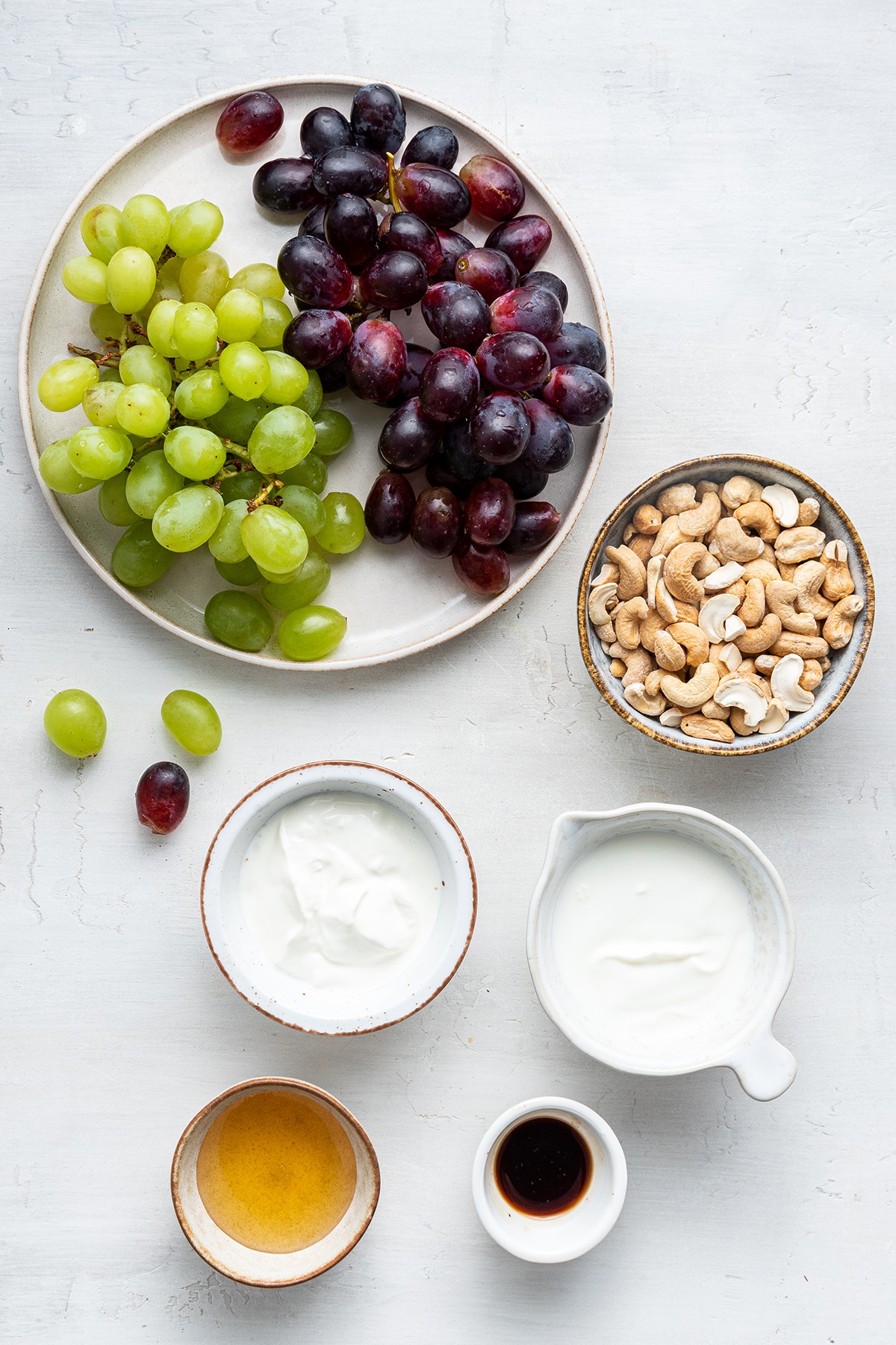 Overhead view of the ingredients needed for grape salad: a plate of red a green grapes, a bowl of cashews, a bowl of Greek yogurt, a bowl of sour cream, a bowl of honey, and a bowl of vanilla extract