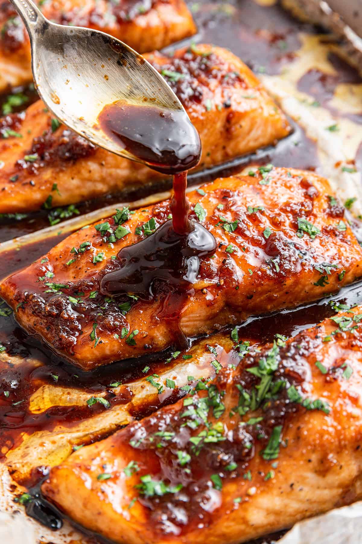 A spoon spooning sauce over honey glazed salmon fillets on a baking sheet