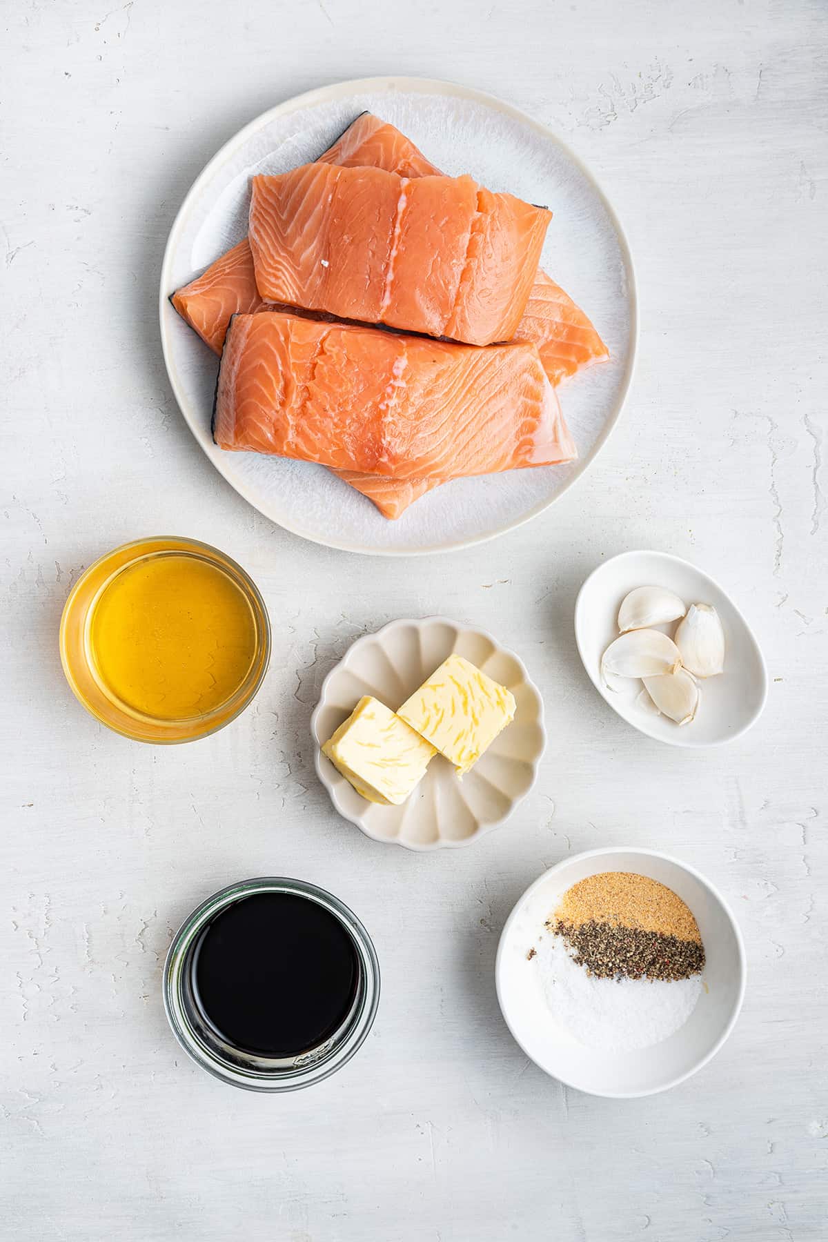 Overhead view of the ingredients needed for honey glazed salmon: a plate of salmon filets, a bowl of soy sauce, a bowl of garlic, a bowl of butter, a bowl of honey, and a bowl of salt, pepper, and garlic powder