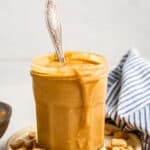 A jar of honey roasted peanut butter on a plate, with a spoon sticking out of it, peanuts on the plate, and a blue and white striped kitchen towel