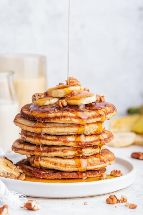Pouring maple syrup onto stack of oatmeal banana pancakes topped with bananas and pecans
