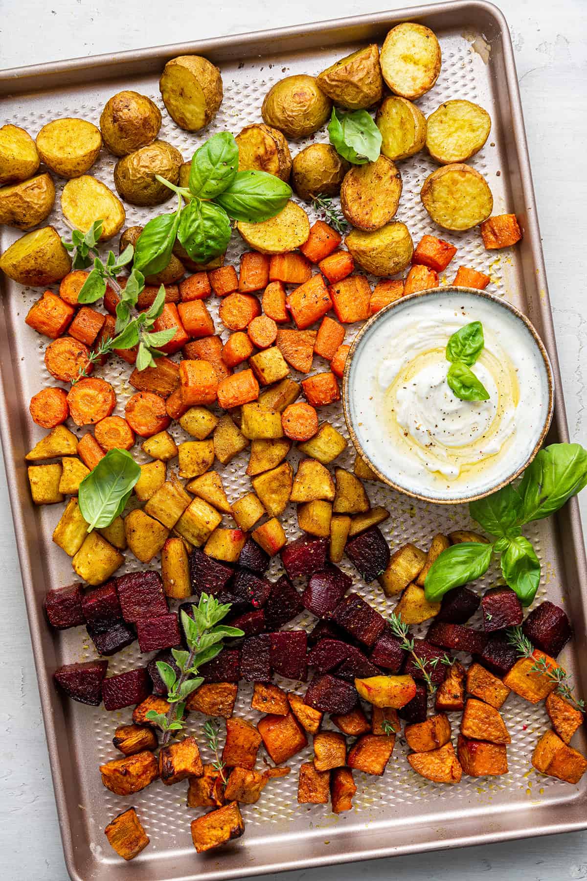 Overhead view of oven-roasted root vegetables on baking dish with fresh herbs and bowl of yogurt dip