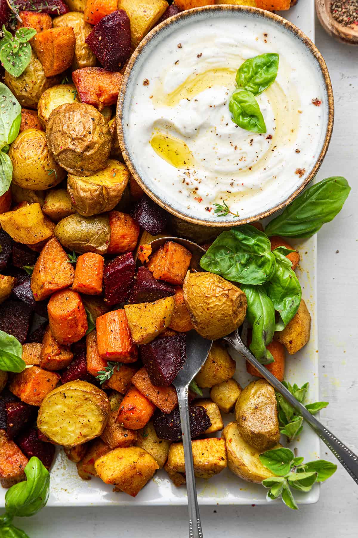 Overhead view of oven-roasted root vegetables on platter with serving spoons and bowl of dip