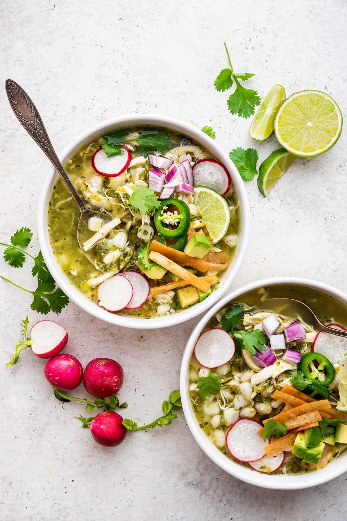 Overhead view of pozole verde in two bowls, garnished with sliced radishes, tortilla strips, lime wedges, and red onions