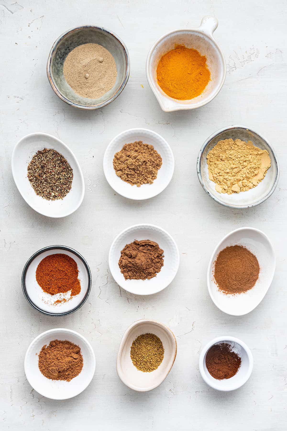 Overhead view of 11 bowls with the ingredients needed for ras el hanout: ground ginger, cardamom, allspice, nutmeg, cinnamon, coriander, turmeric, black pepper, cayenne pepper, ground anise, and ground cloves