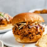 Vegan lentil sloppy joe sandwich on a hamburger bun with tortilla chips and vegetable slaw on a white plate with a stripped dishcloth