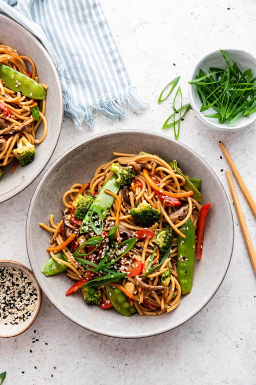 Overhead view of two bowls of vegetable lo mein with small bowls of sliced green onions and sesame seeds