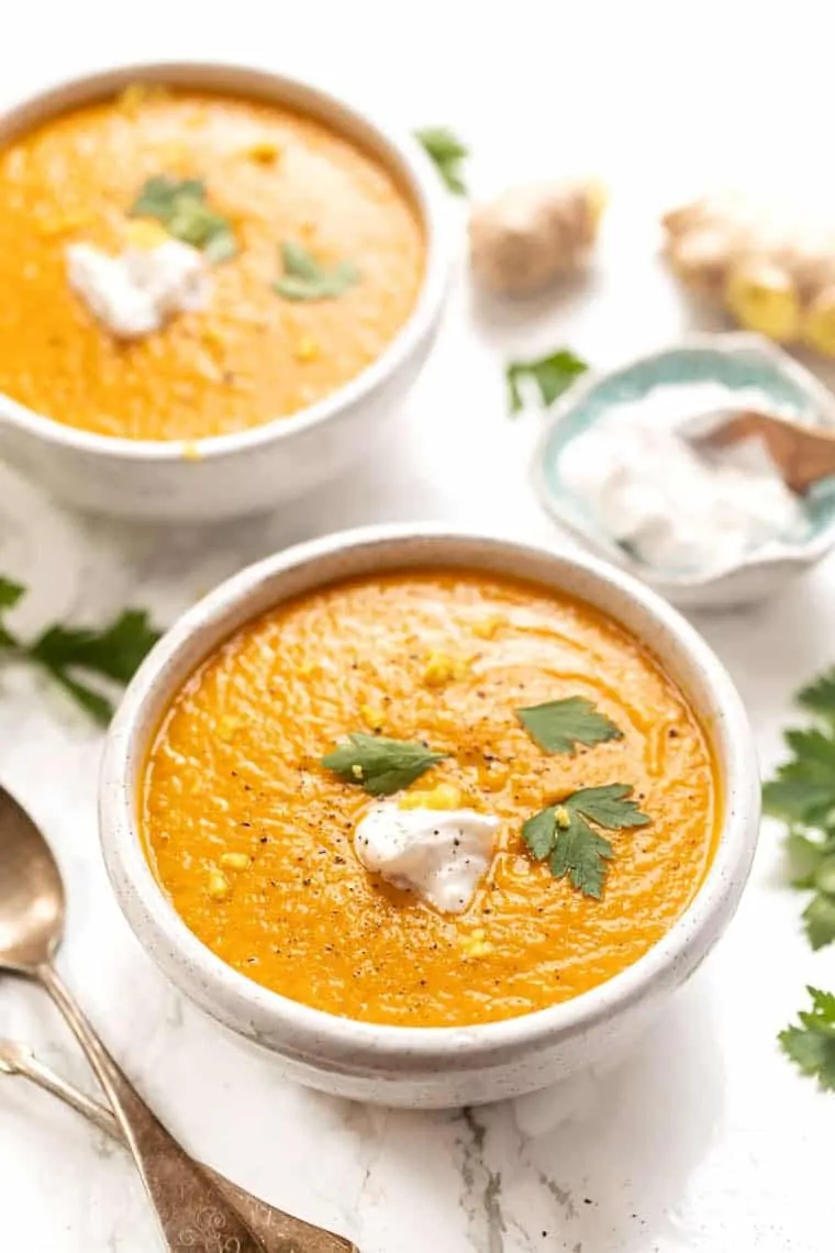 A bowl of turmeric carrot soup, topped with vegan coconut yogurt and three parsley leaves, with another bowl of soup and a bowl of coconut yogurt in the background, surrounded by spoons and parsley