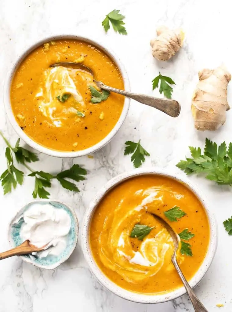 Overhead view of two bowls of turmeric carrot soup topped with vegan coconut yogurt and parsley, with spoons in the bowl, next to a bowl of vegan coconut yogurt, fresh parsley, and fresh ginger. 