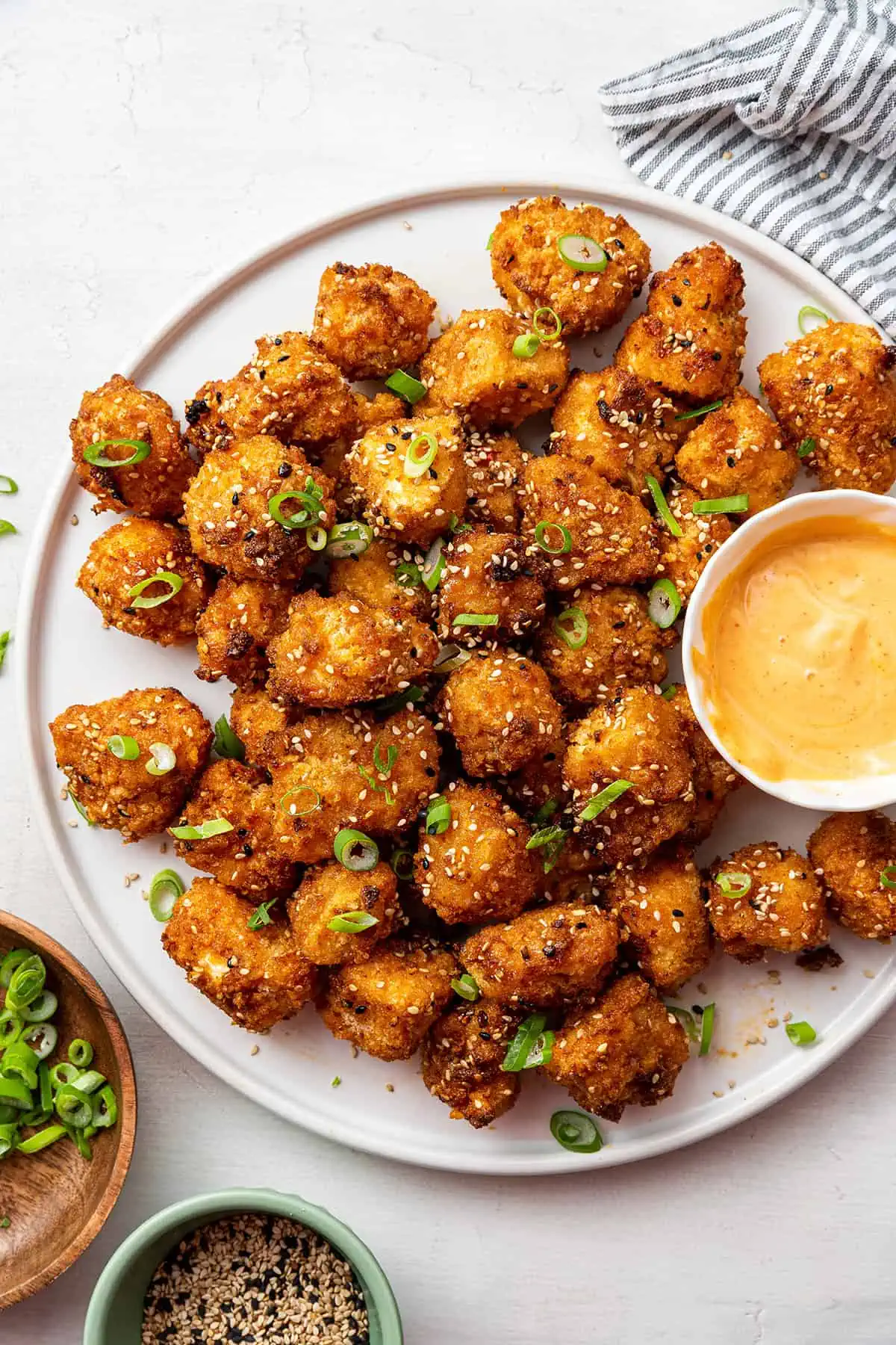 Overhead view of a plate of bang bang cauliflower topped with scallions and sesame seeds and a bowl of sauce