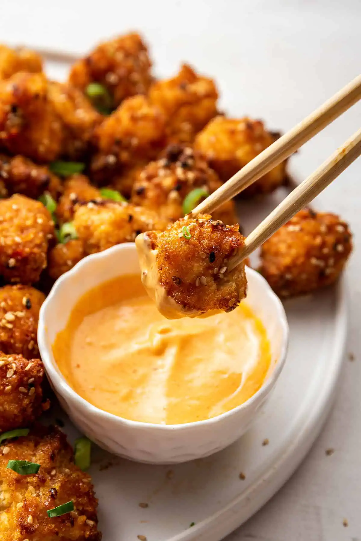 A pair of chopsticks dipping a piece of bang bang cauliflower into dipping sauce, with a serving tray of cailflower behind it