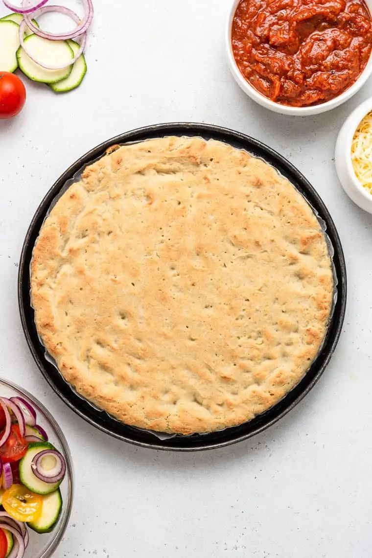 overhead of baked gluten-free pizza crust on a pan with sauce in a bowl on the side