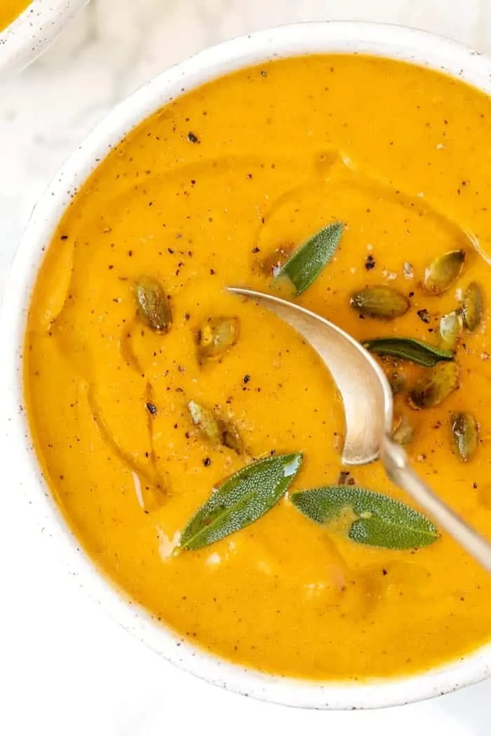 Close up of a spoon dipping into a bowl of roasted pumpkin soup, garnished with pumpkin seeds and whole sage leaves