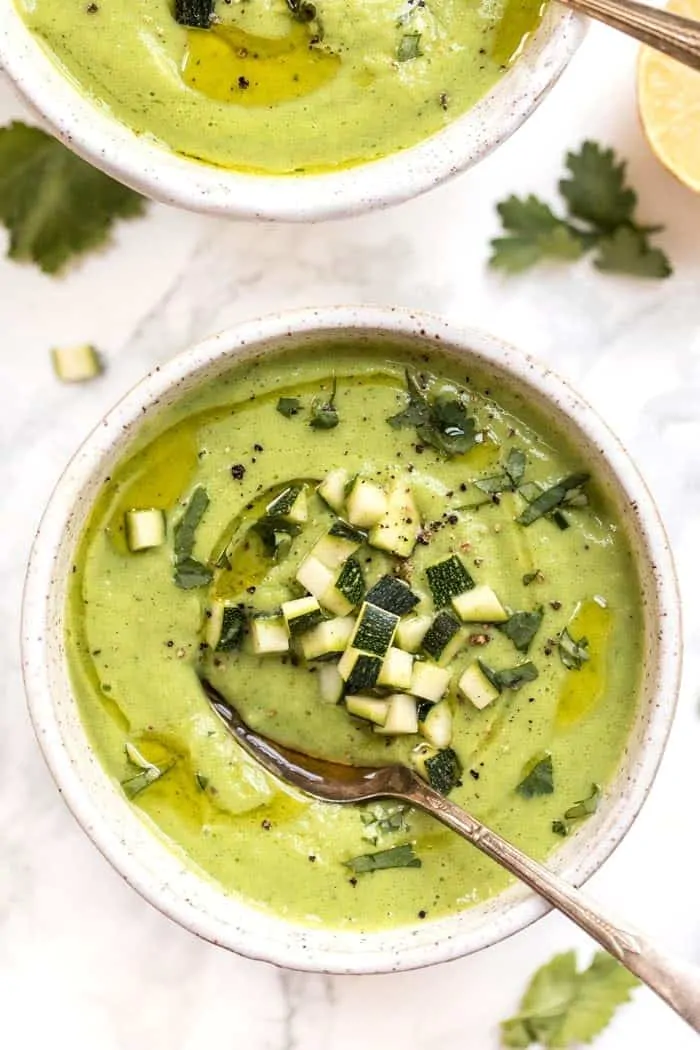 A bowl of avocado and zucchini soup with cubes of zucchini on top, and a spoon in it