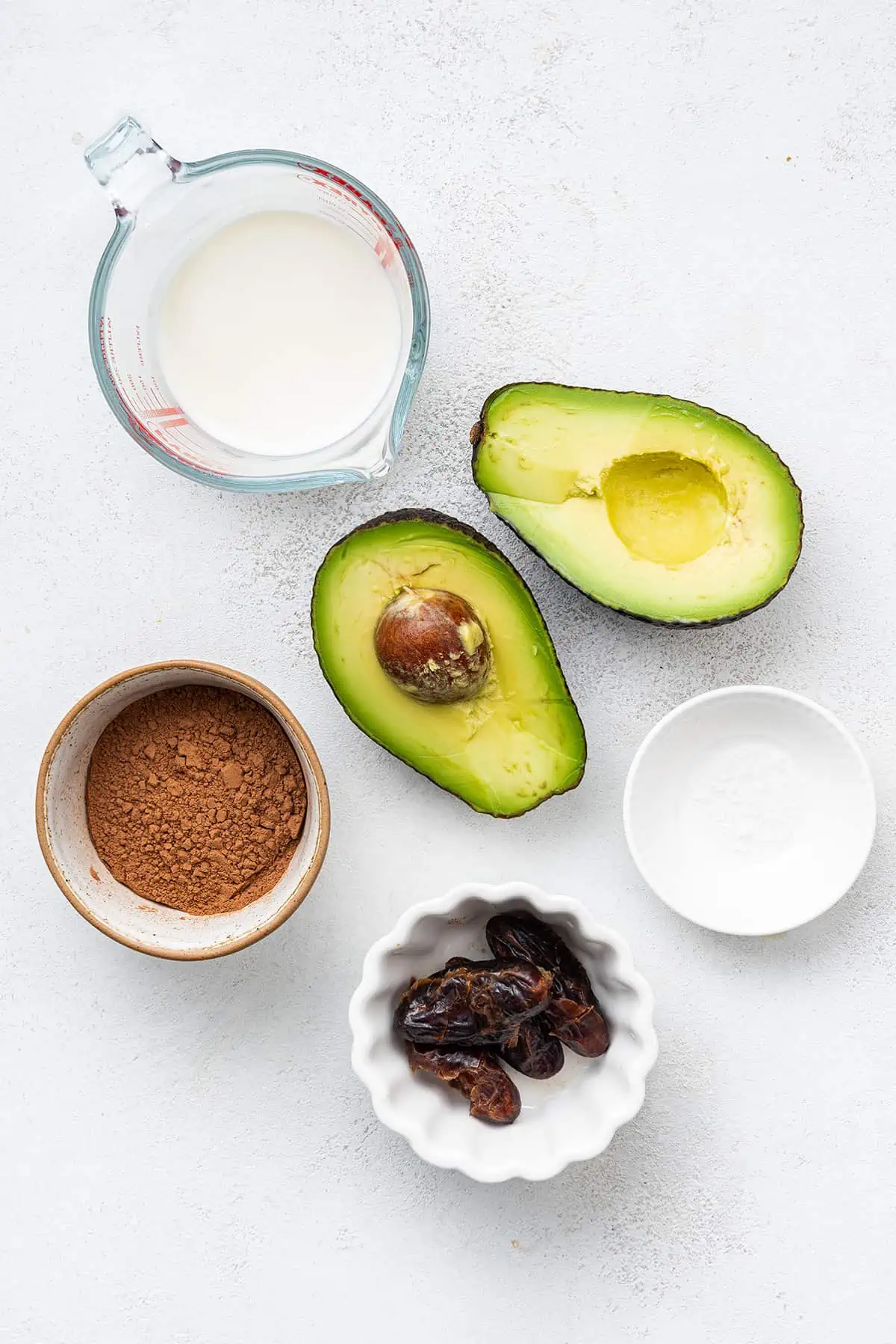Overhead view of the ingredients needed for dark chocolate avocado mousse: an avocado cut in half, a bowl of dates, a bowl of cacao powder, a bowl of vegan milk, and a bowl of salt
