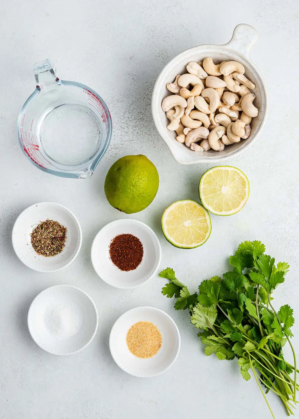 Overhead view of the ingredients needed for cilantro lime sauce: a bowl of cashews, a pyrex of water, a bowl of salt, a bowl of pepper, a bowl of chili powder, a bowl of garlic powder, a bunch of cilantro, a whole lime, and a lime cut in half