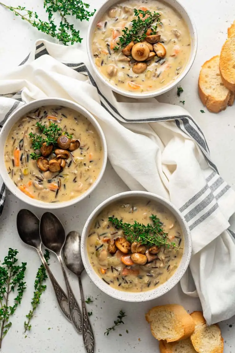 Overhead view of three bowls of wild rice soup topped with sautéed mushrooms and thyme sprigs, surrounded by spoons, thyme sprigs, slices of bread, and kitchen towels.
