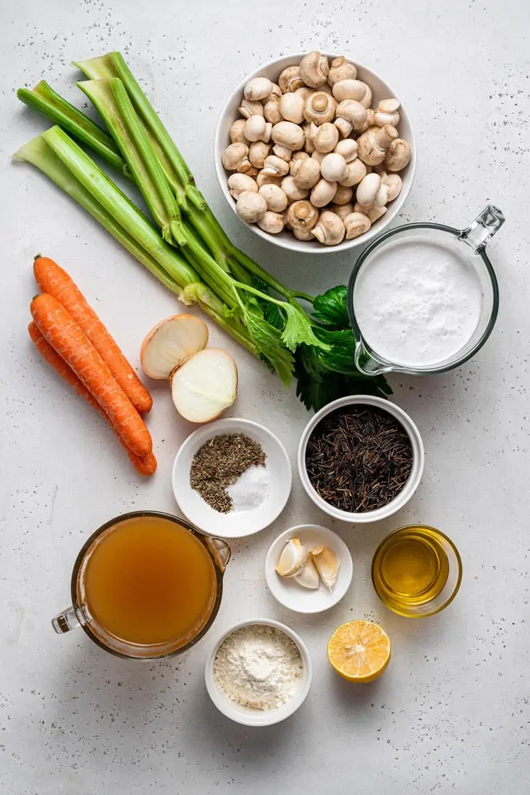 Overhead view of the ingredients needed for creamy mushroom and wild rice soup: an onion cut in half, three carrots, four stalks of celery, a bowl of mushrooms, a pyrex of coconut milk, a bowl of salt, pepper, and Italian seasoning, a bowl of wild rice, a bowl with three garlic cloves, a pyrex of veggie broth, a bowl of flour, a glass of oil, and half a lemon