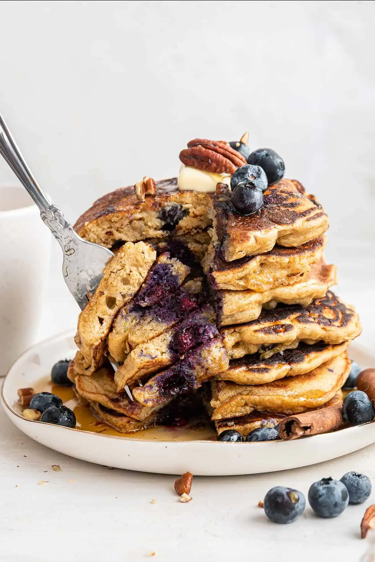 A fork taking a large bite out of a stack of seven blueberry pancakes on a white plate, topped with butter, blueberries, and a pecan