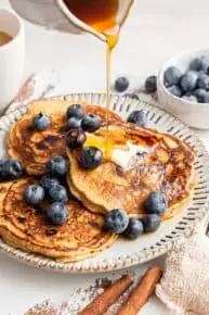 Maple syrup being poured onto a plate of three pancakes topped with blueberries and butter, with blueberries behind them