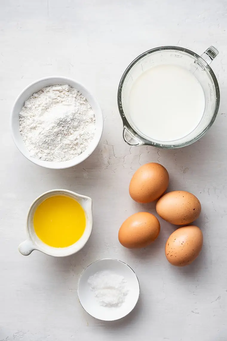 All the ingredients for gluten-free popovers: a bowl of milk, a pourer of melted butter, a bowl of salt, a bowl of gluten-free all-purpose flour blend, and four raw eggs
