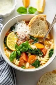 Overhead view of vegetable stew in bowl with Parmesan, lemon slice, basil, and bread