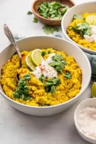 A bowl of kitchari topped with yogurt, cilantro, and limes, with a spoon in it, with a bowl of kitchari and a bowl of cilantro in the background