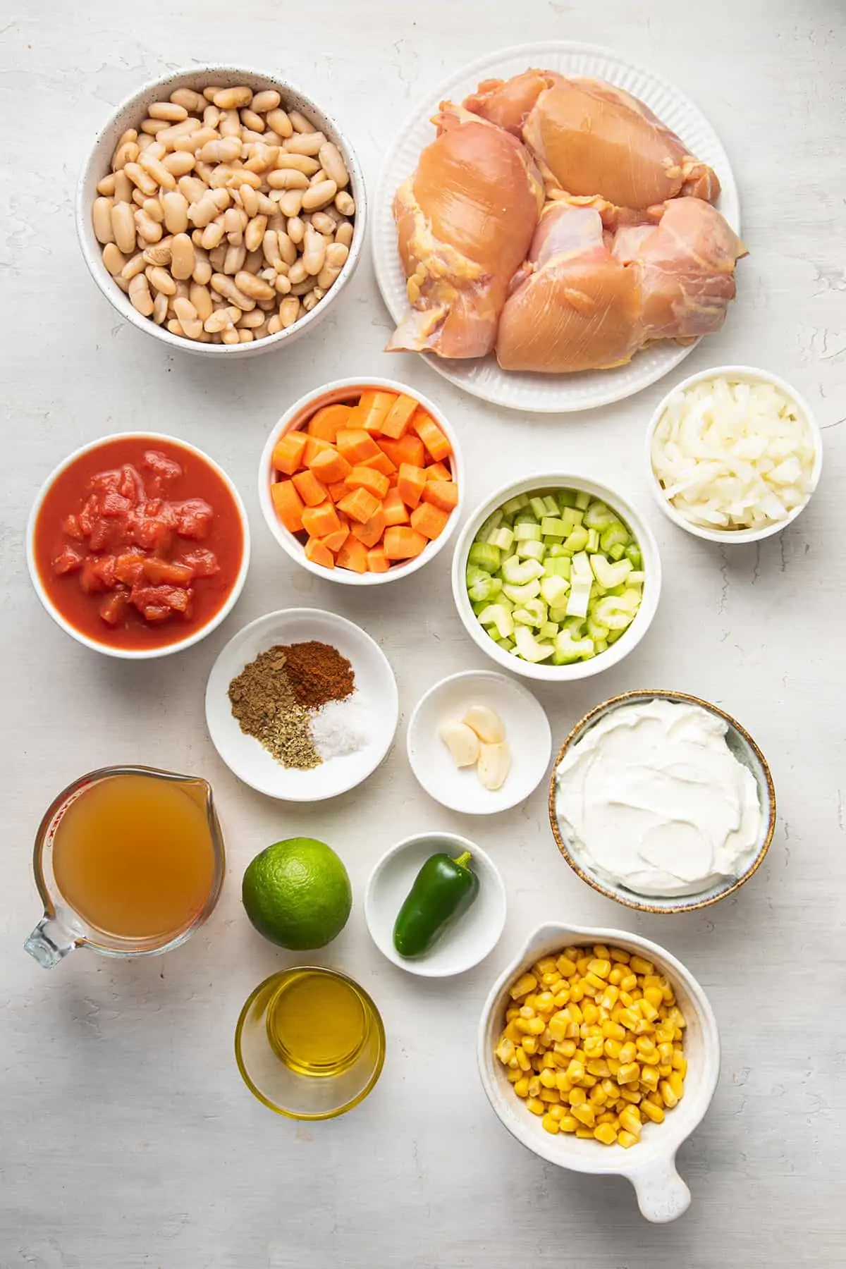 Overhead picture of the ingredients for white chicken chili: beans, chicken thighs, canned tomatoes, carrots, celery, onion, spices, garlic, sour cream, vegetable broth, a lime, a jalapeño, oil, and corn