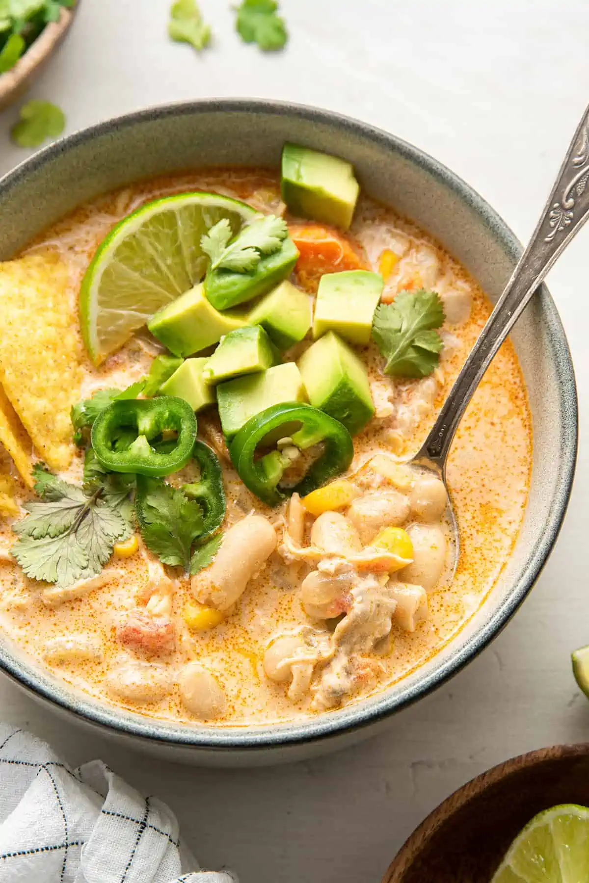 A bowl of white chicken chili garnished with chilis, cilantro, avocado, and a lime spice, with a spoon in the bowl