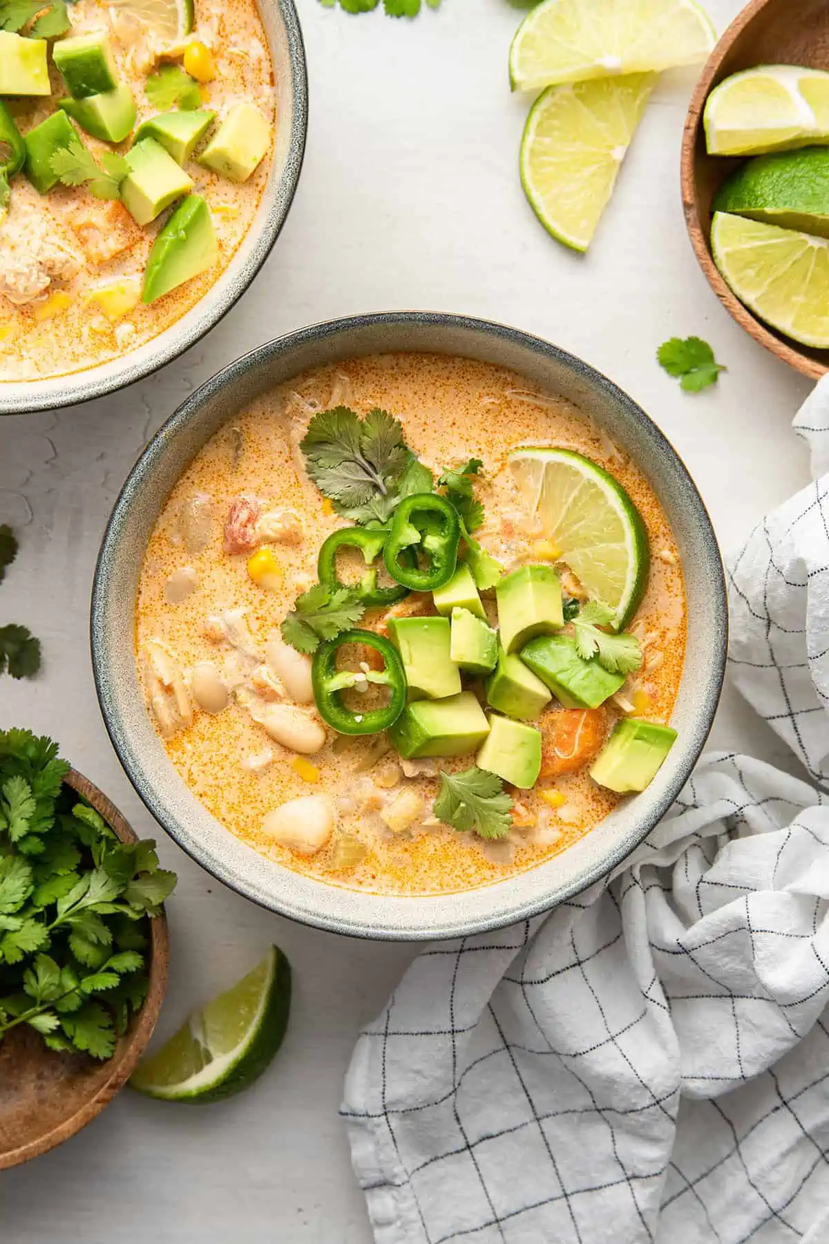 Overhead view of a bowl of white chicken chili topped with limi, chilis, avocado, and cilantro, next to another bowl of chili, a bowl of cilantro, and lime wedges