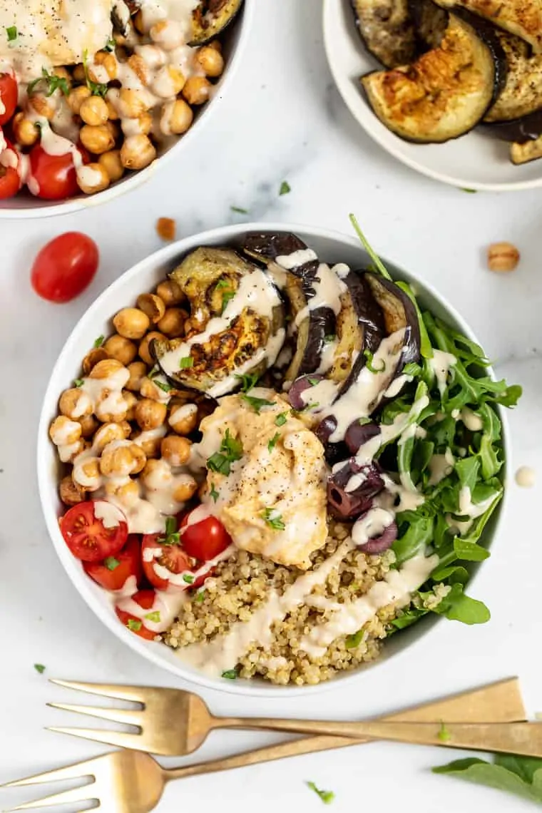 Overhead view of a quinoa bowl topped with hummus, arugula, olives, tomatoes, chickpeas, and eggplant, next to two forks 