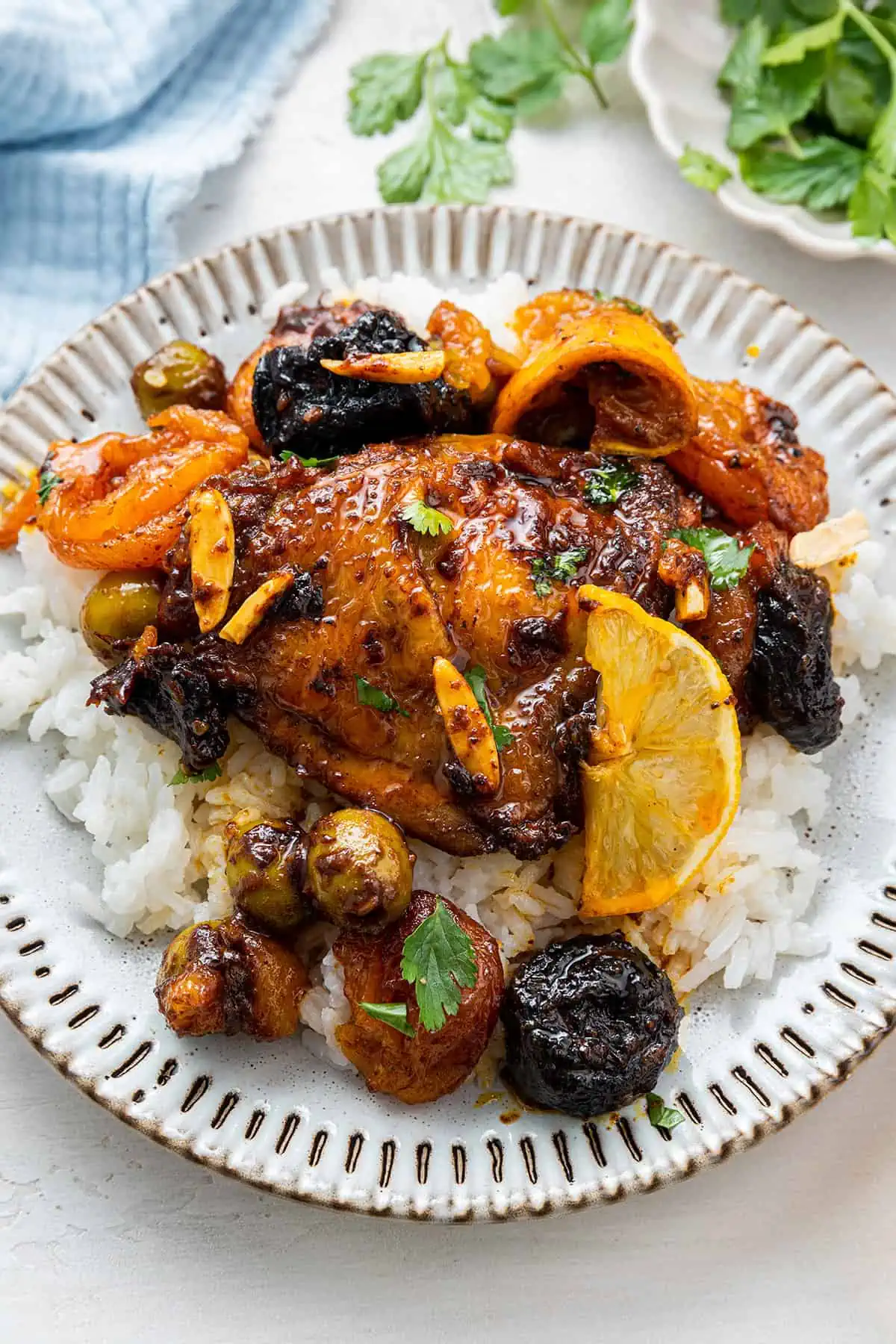Overhead view of a plate with a chicken thigh on top of rice, covered with lemon slices, slivered almonds, prunes, olives, and dried apricots