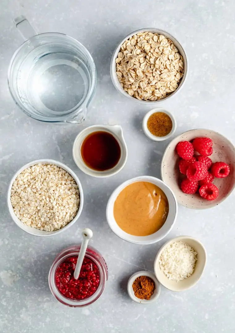 overhead of ingredients to make oatmeal with peanut butter, jam, water, berries, coconut, maple syrup and cinnamon