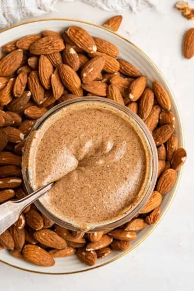Overhead view of a jar of pumpkin spice almond butter with a spoon submerged in it, sitting on a plate covered in almonds