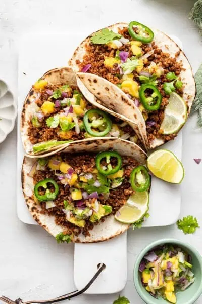 Three vegan tacos on white board with garnishes and lime wedges