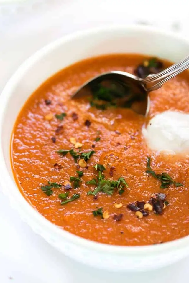 Close up of a bowl of roasted red pepper soup garnished with parsley, sour cream, and chili flakes, with a spoon in it