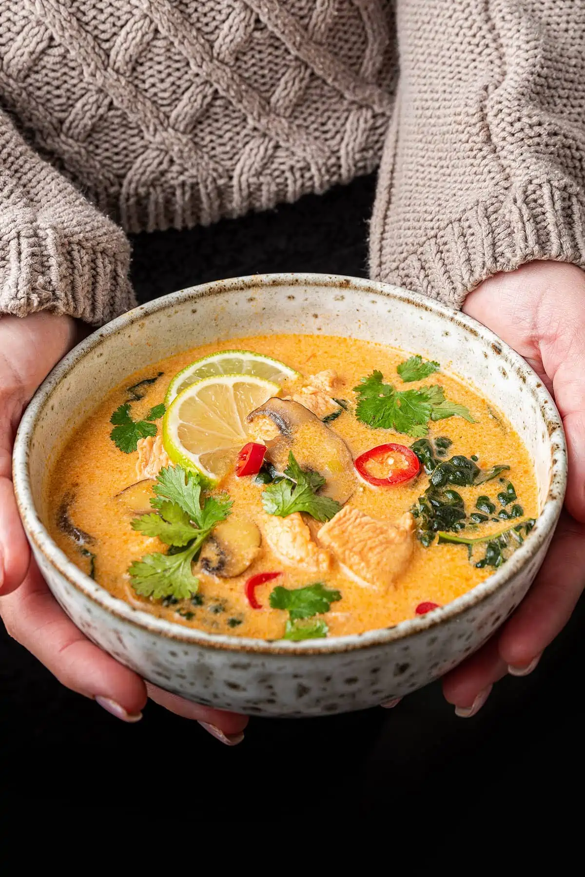A bowl of Thai coconut curry sou, topped with chilis, limes, and cilantro, being held in two hands.