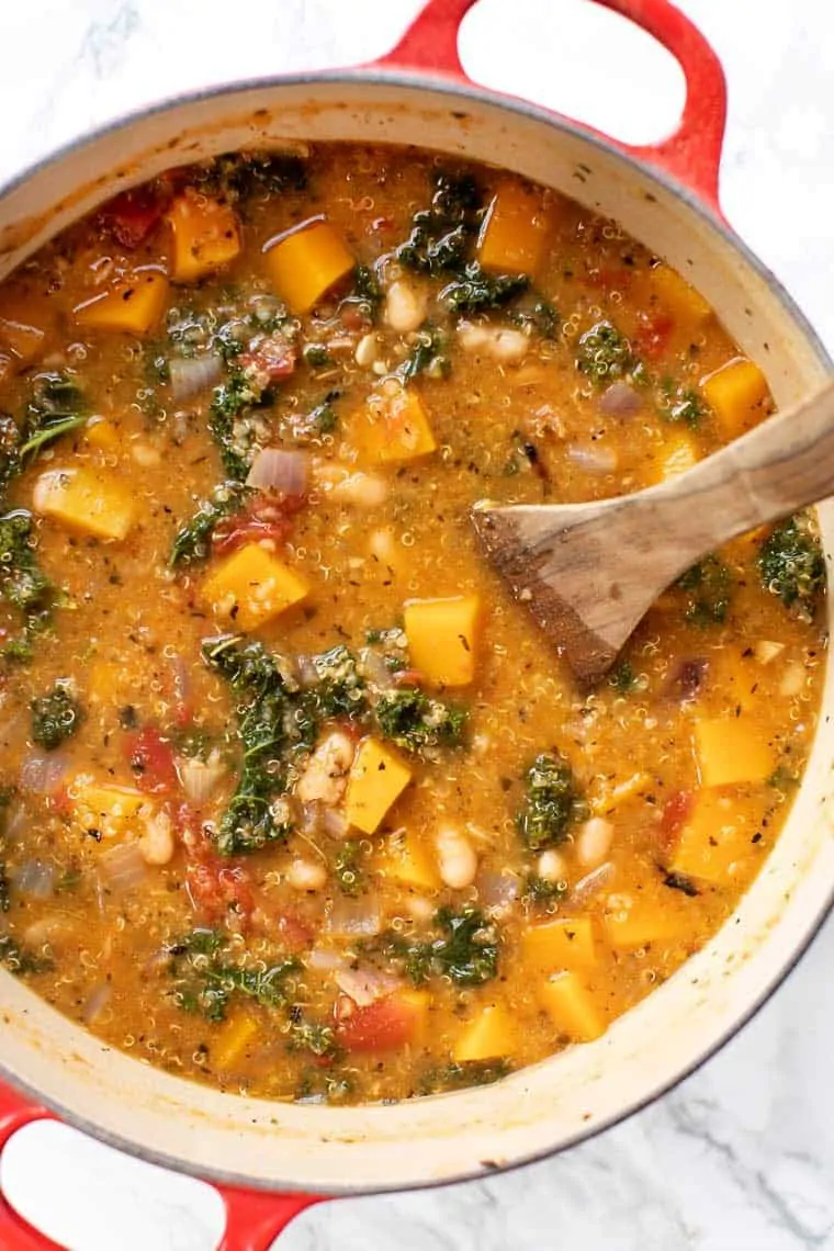 Overhead view of soup cooking in a Dutch oven with a wooden spoon in it, with pieces of squash, kale, and white beans