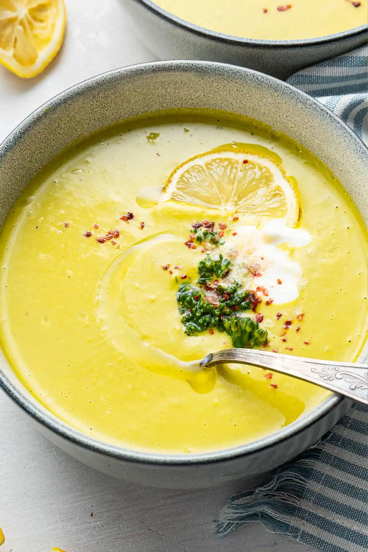 A bowl of broccoli soup garnished with broccoli, chili flakes, vegan creme fraiche, and a lemon slice, with a spoon in it